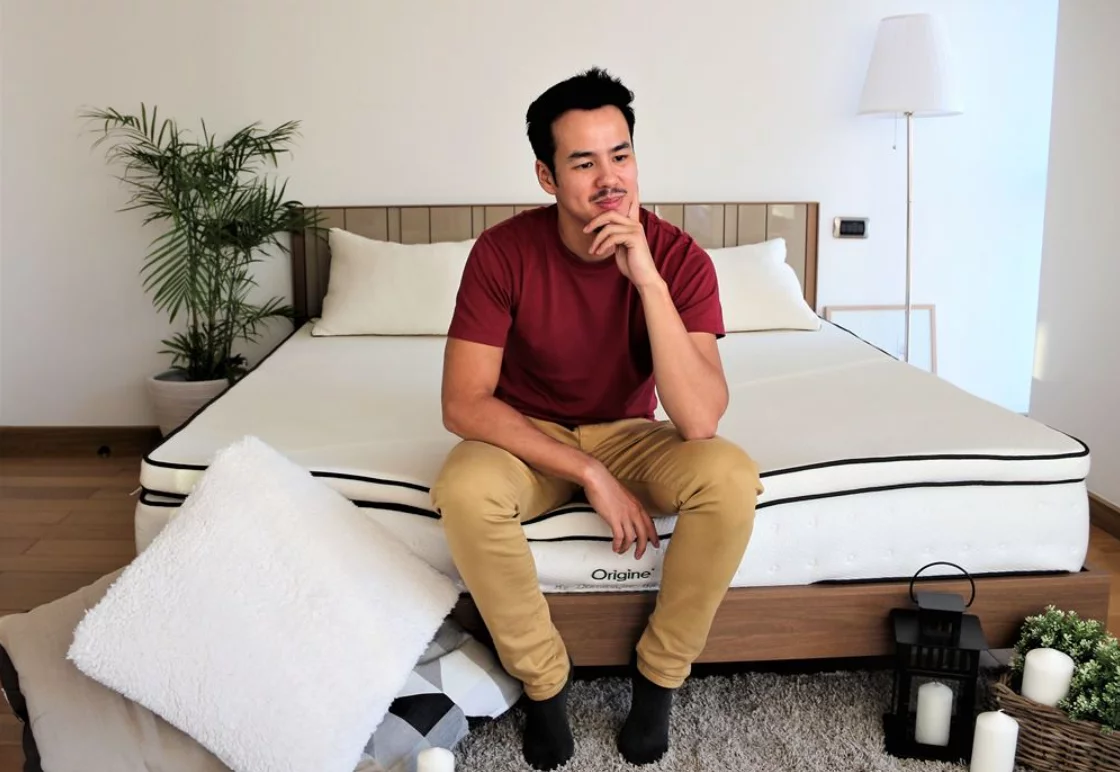 Un jeune homme est assis sur un matelas en latex Origine, l'air pensif. Il porte une chemise rouge et un pantalon kaki et est entouré d'oreillers en latex.