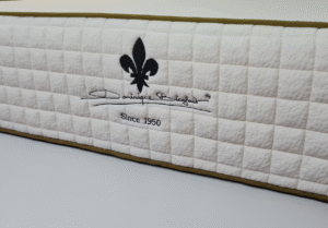 Close-up of a premium mattress side panel with quilted texture, featuring a black fleur-de-lis logo, the signature 'Dominique Bienfait,' and the inscription 'Since 1950.