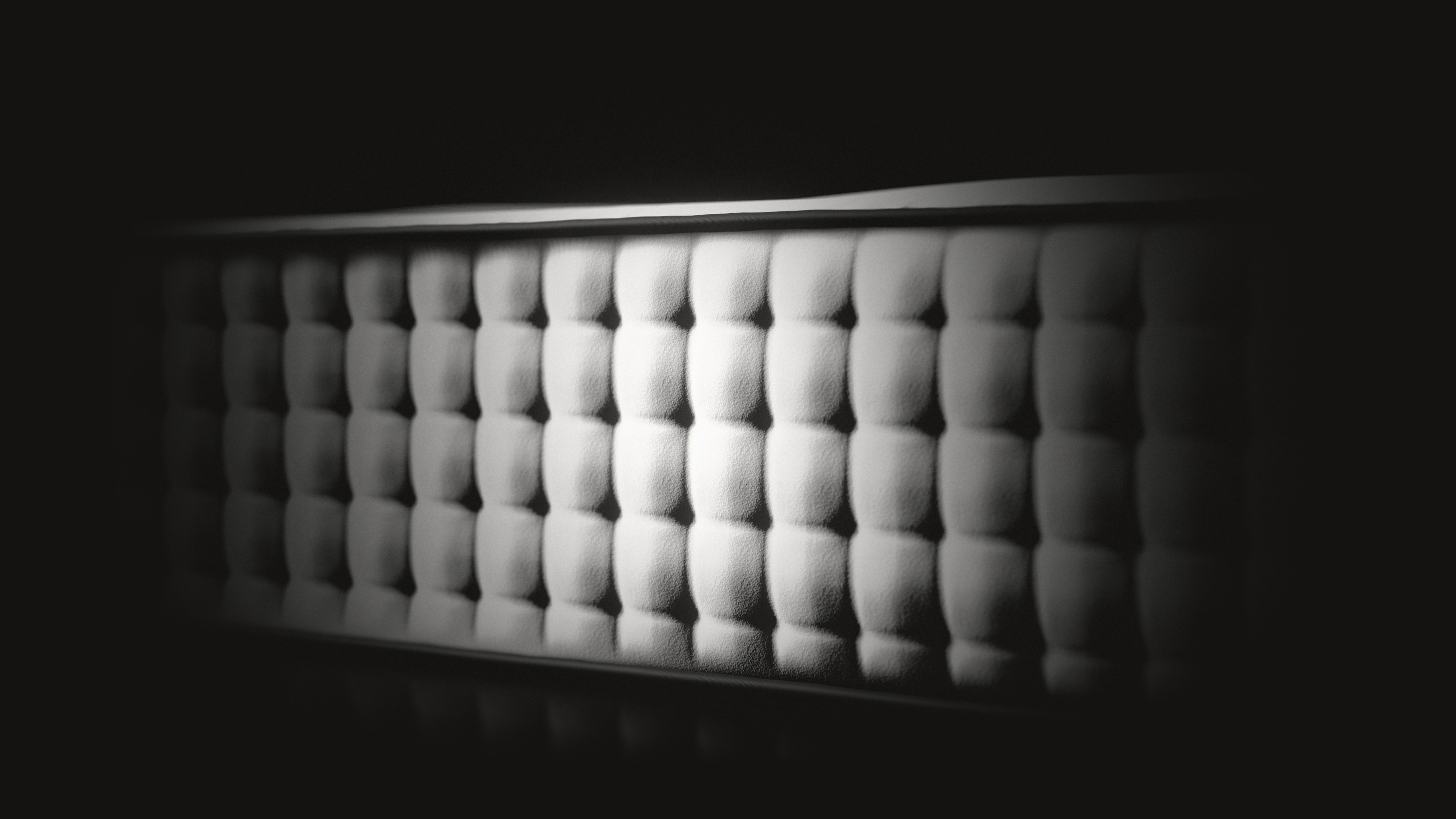 The side of the mattress, close, in low raking light: rows of soft white cushions with deep buttons between them, under a black cord, fading into the dark.