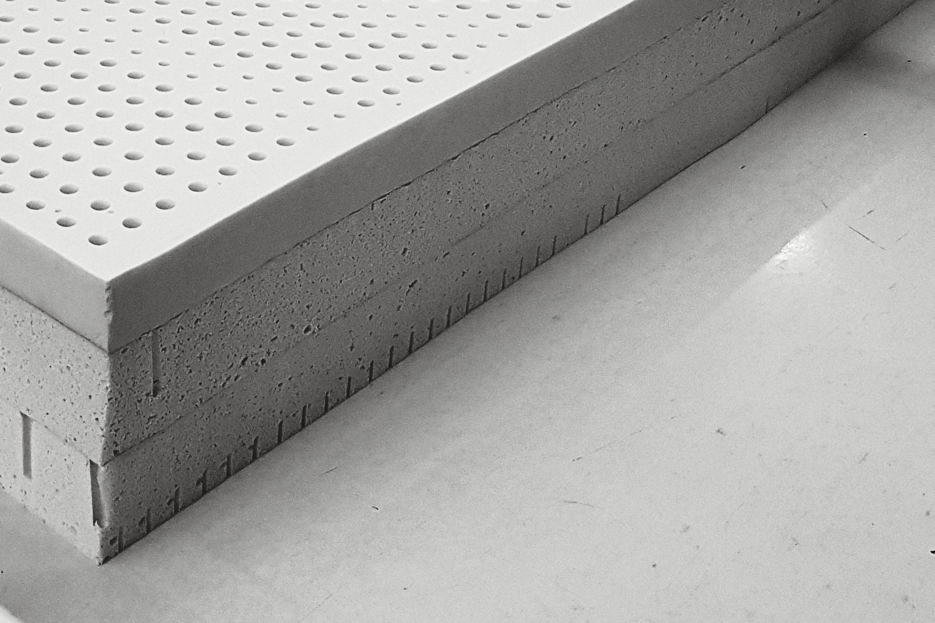 The near corner of a stack of three slabs of latex on a white table: the smooth perforated top, the open cells of the cut sides, a row of slits along the lower edge.