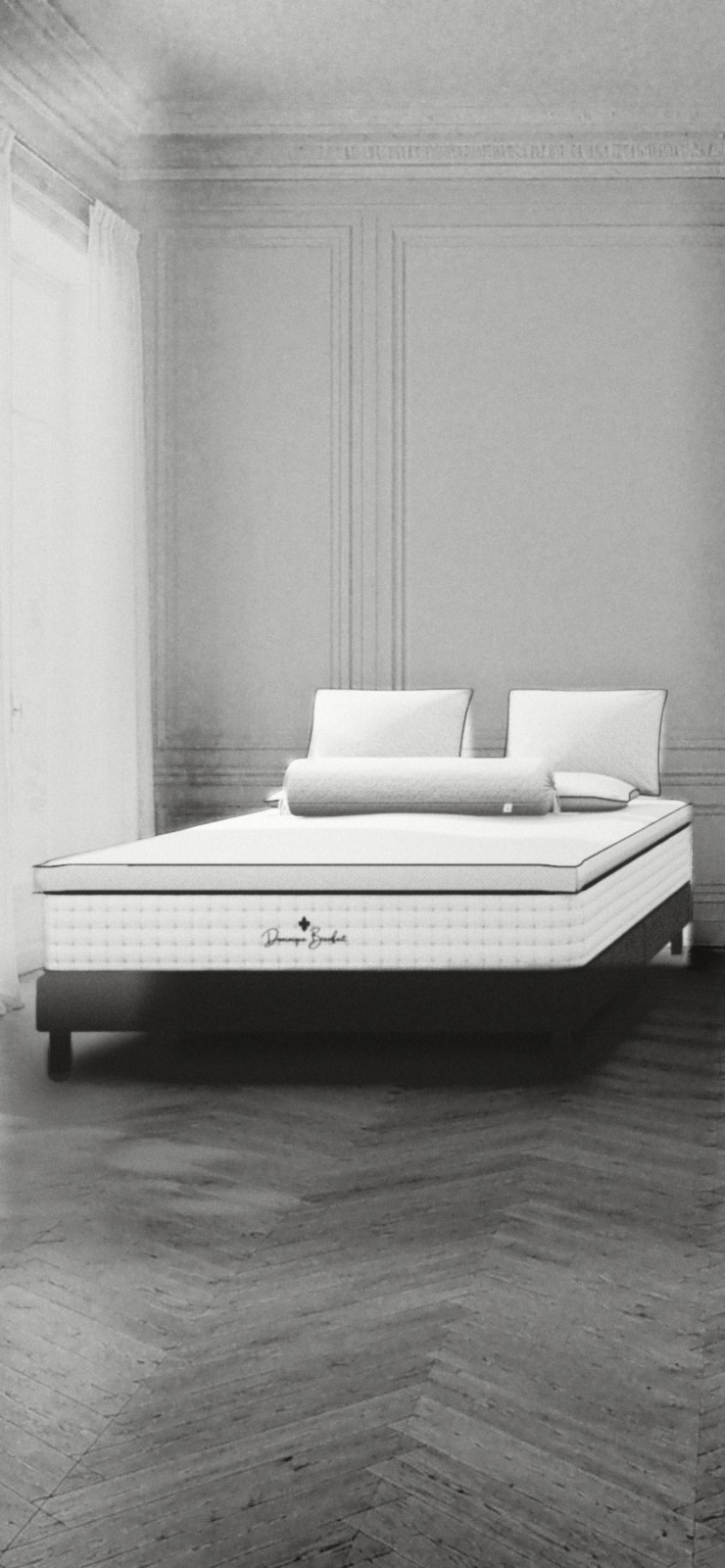 A bed in a panelled room with tall curtained windows and a herringbone parquet: a white mattress with tufted sides on a dark base, a topper turned back at its foot, four pillows. On the foot end, a black fleur-de-lis and the handwritten name Dominique Bienfait.