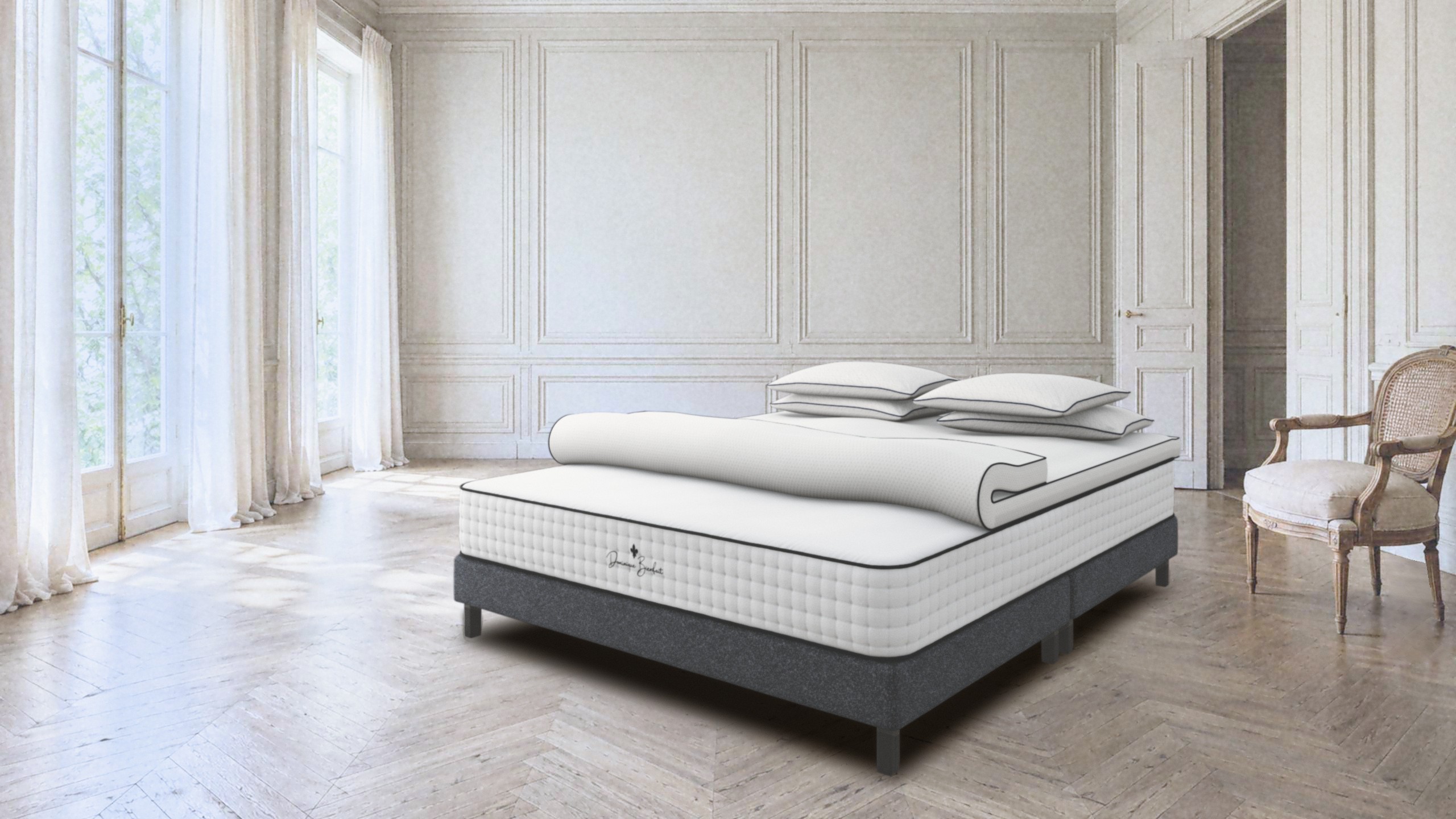 A bed in a bright panelled room with tall windows and pale parquet: a white mattress with tufted sides on a charcoal base, a topper turned back at its foot, four pillows. On the foot end, a black fleur-de-lis and the handwritten name Dominique Bienfait.