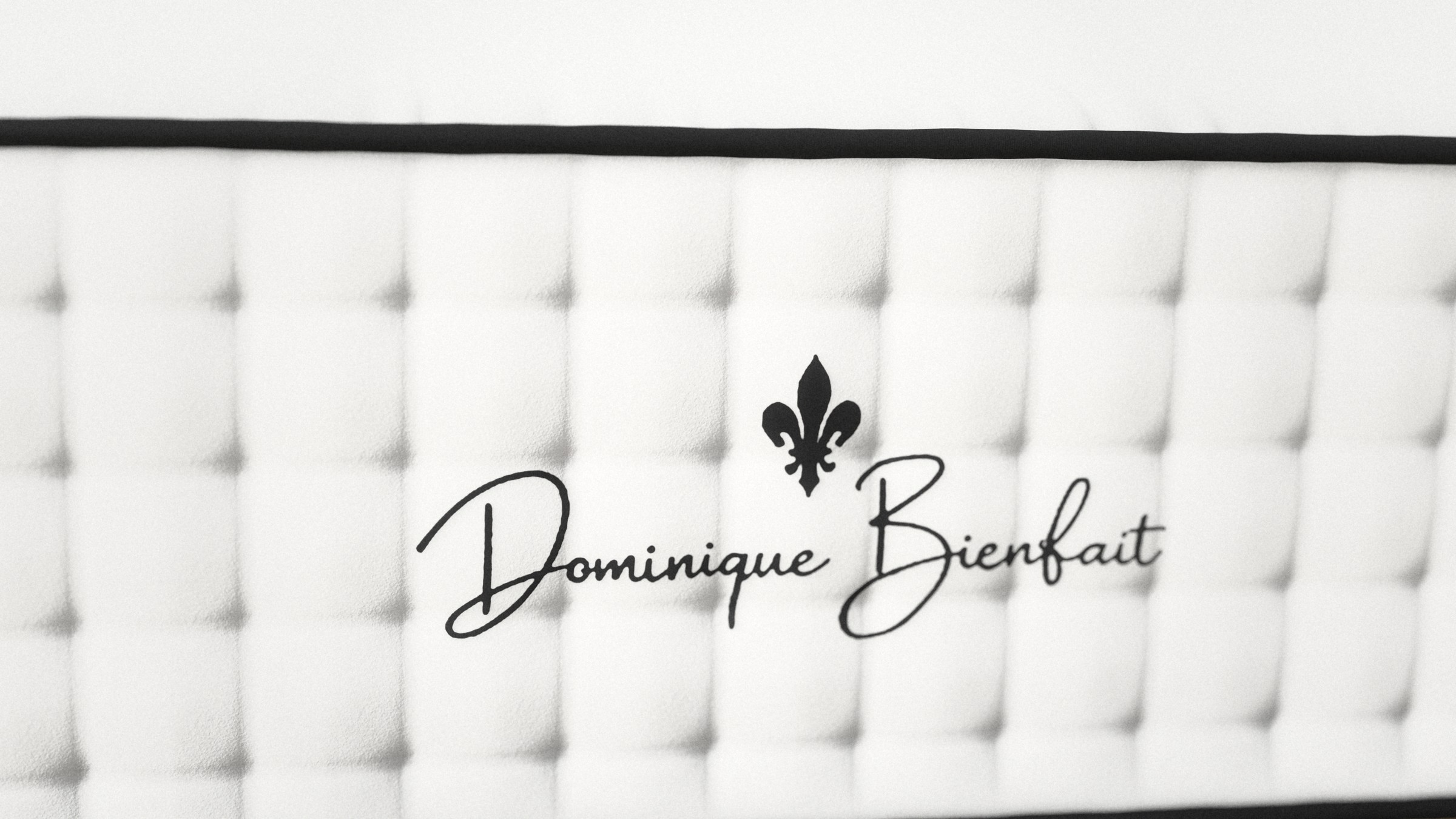 ด้านปลายเท้าของที่นอนในระยะใกล้: สัญลักษณ์เฟลอร์เดอลิสสีดำและชื่อ Dominique Bienfait บนแผงสีขาวแบบดึงกระดุม ใต้เชือกสีดำ