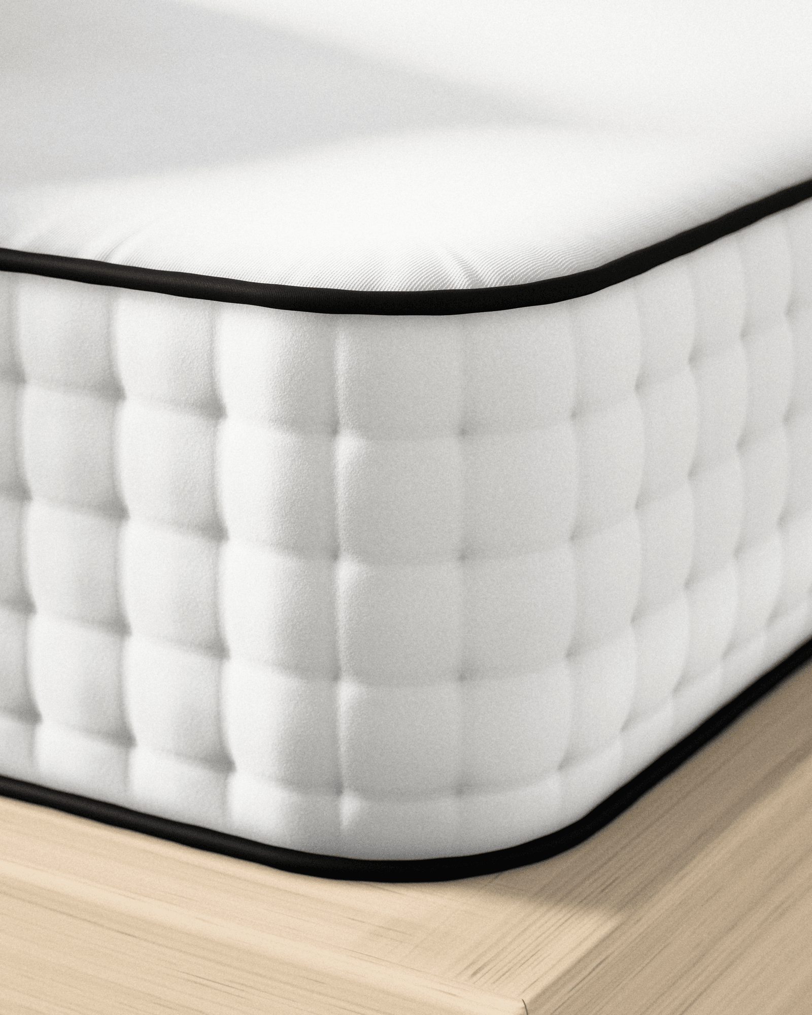The corner of the mattress on a pale oak base: the white cotton top rolls into a black cord, and the tufted side falls away below it in low sunlight.