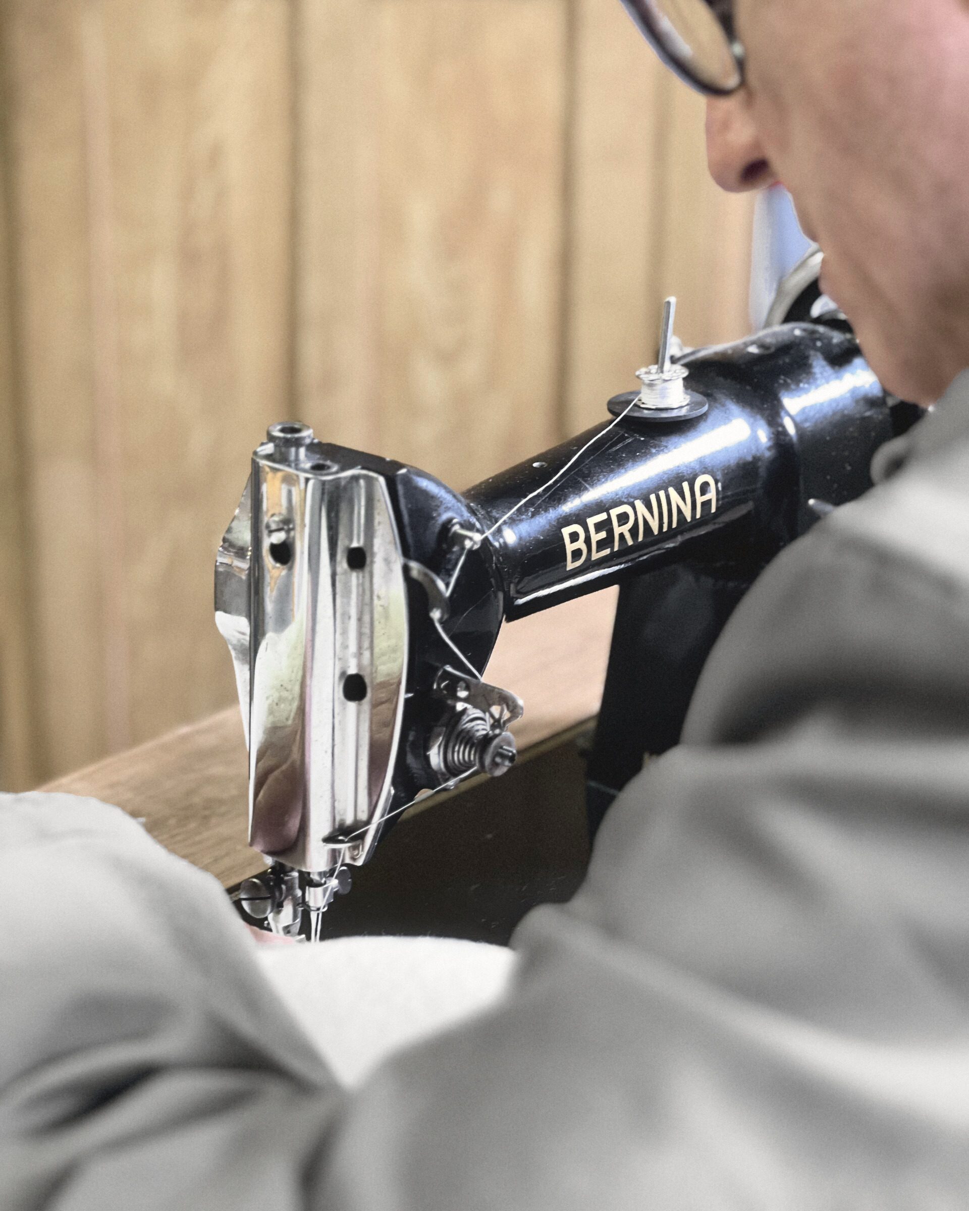 มองข้ามไหล่: คนหนึ่งนั่งอยู่ที่จักรเย็บผ้า Bernina รุ่นเก่าสีดำ ผ้าสีขาวอยู่ใต้เข็ม และประตูไม้อยู่ด้านหลัง