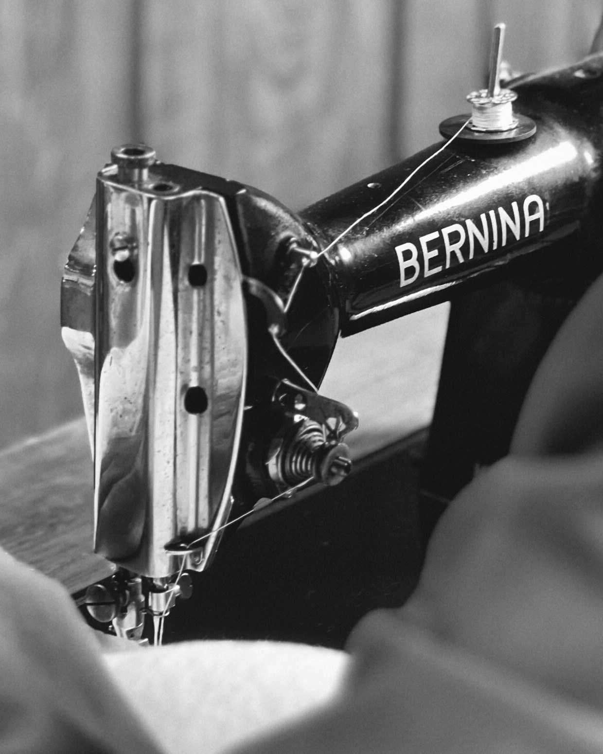 The head of an old black Bernina sewing machine, thread running from the bobbin down to the needle, a shoulder out of focus in front, a wooden door behind.