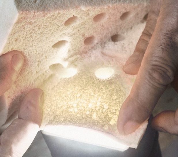 Two hands bending a piece of latex over a lamp, the light showing its open cells round the pin holes.