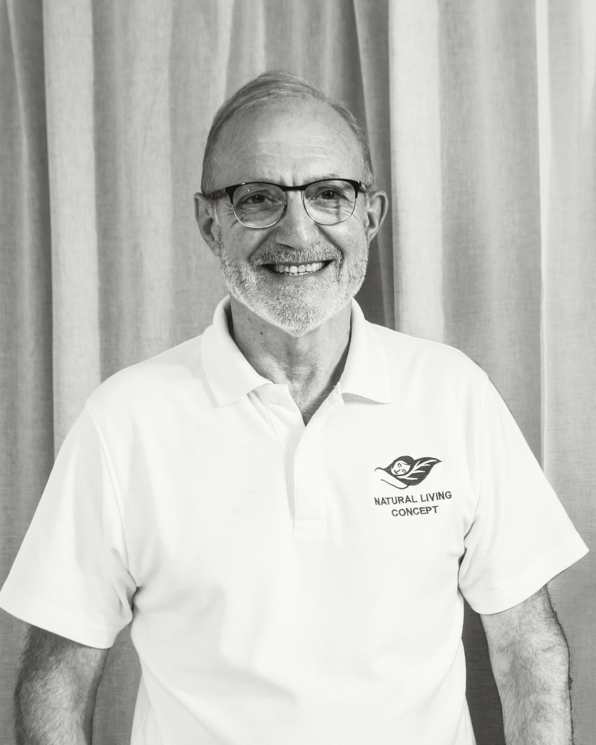 Dominique Bienfait, smiling, in glasses and a white Natural Living Concept polo, against a white curtain.