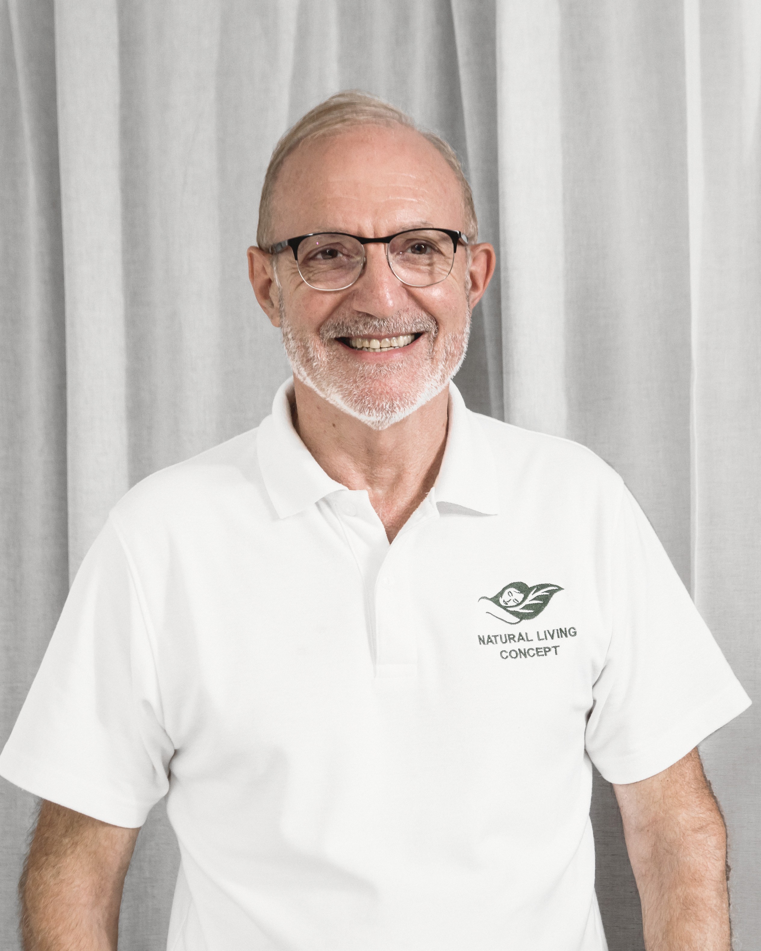 Dominique Bienfait, smiling, in a white polo shirt embroidered with the Natural Living Concept leaf, standing in front of a pale curtain.