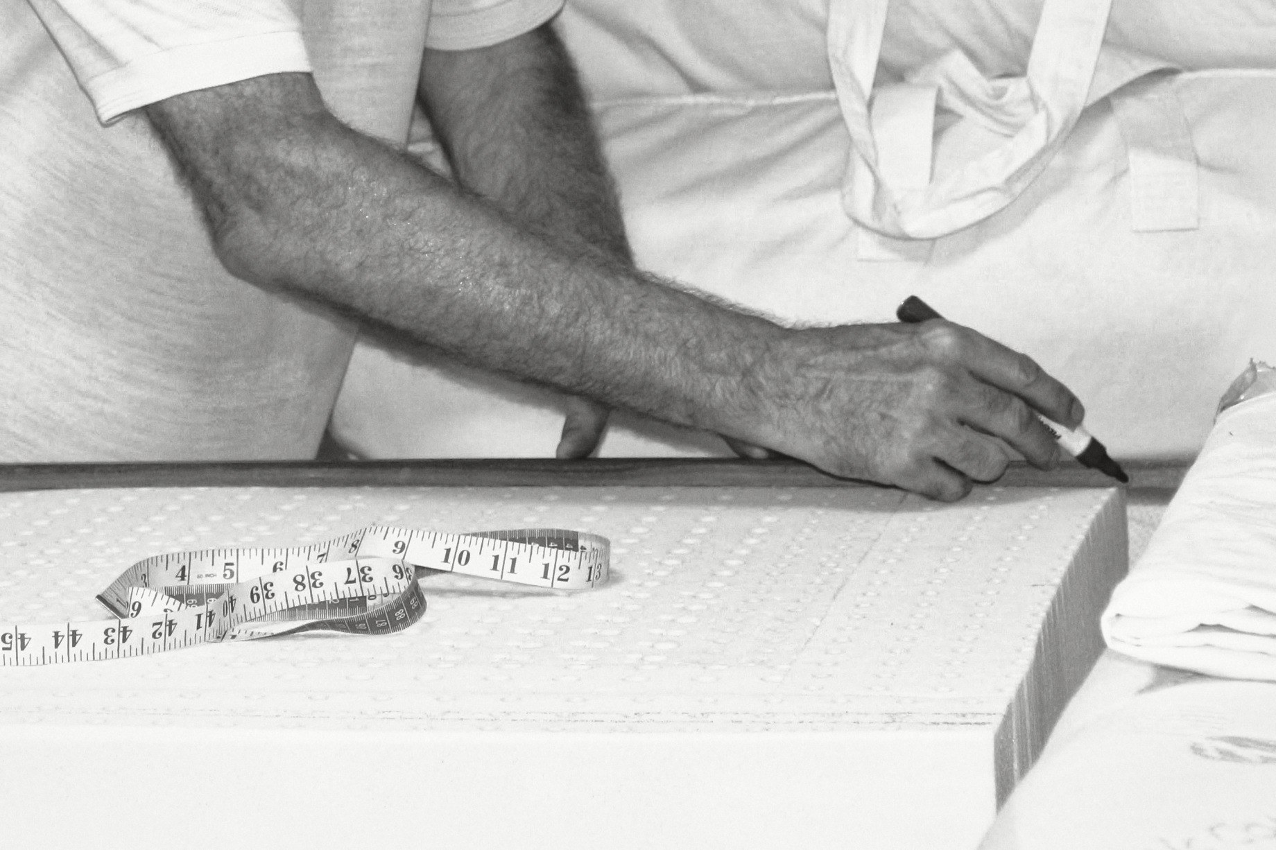 A hand holding a marker against a wooden rule on a block of perforated latex, the forearm reaching in from the left, a tape measure lying on the block.