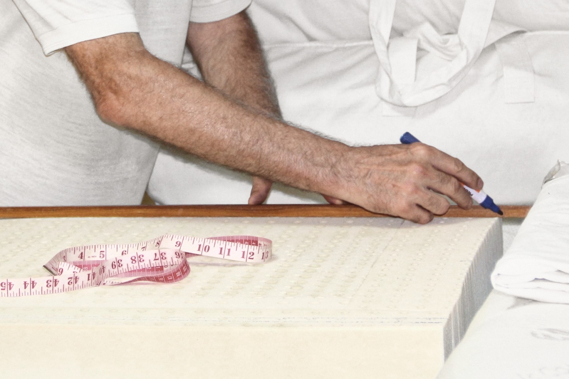 A forearm and a hand drawing a line with a marker along a wooden rule laid across a block of perforated latex, a tape measure lying on the block, white cotton covers behind.