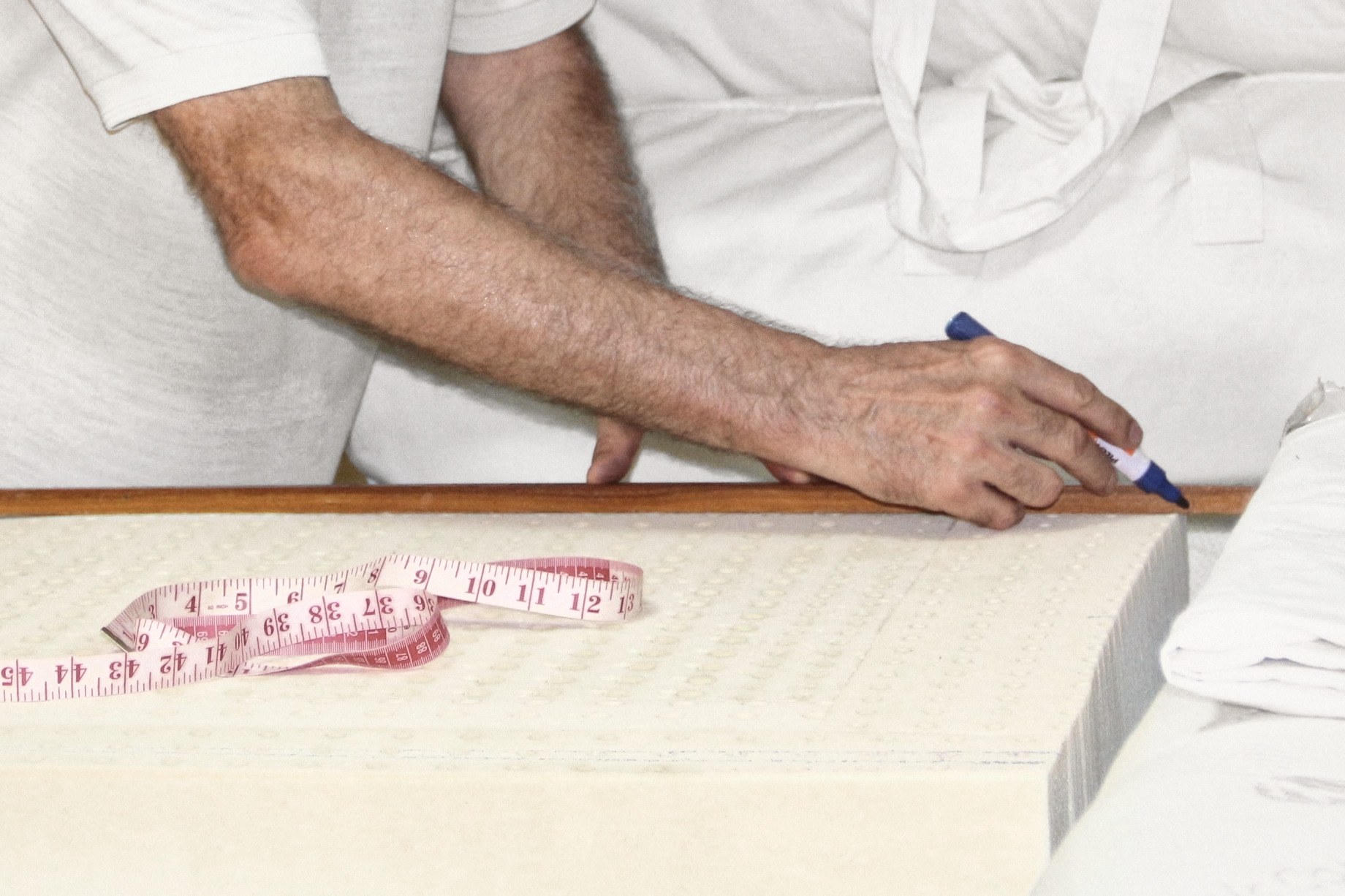 A hand holding a marker against a wooden rule on a block of perforated latex, the forearm reaching in from the left, a tape measure lying on the block.