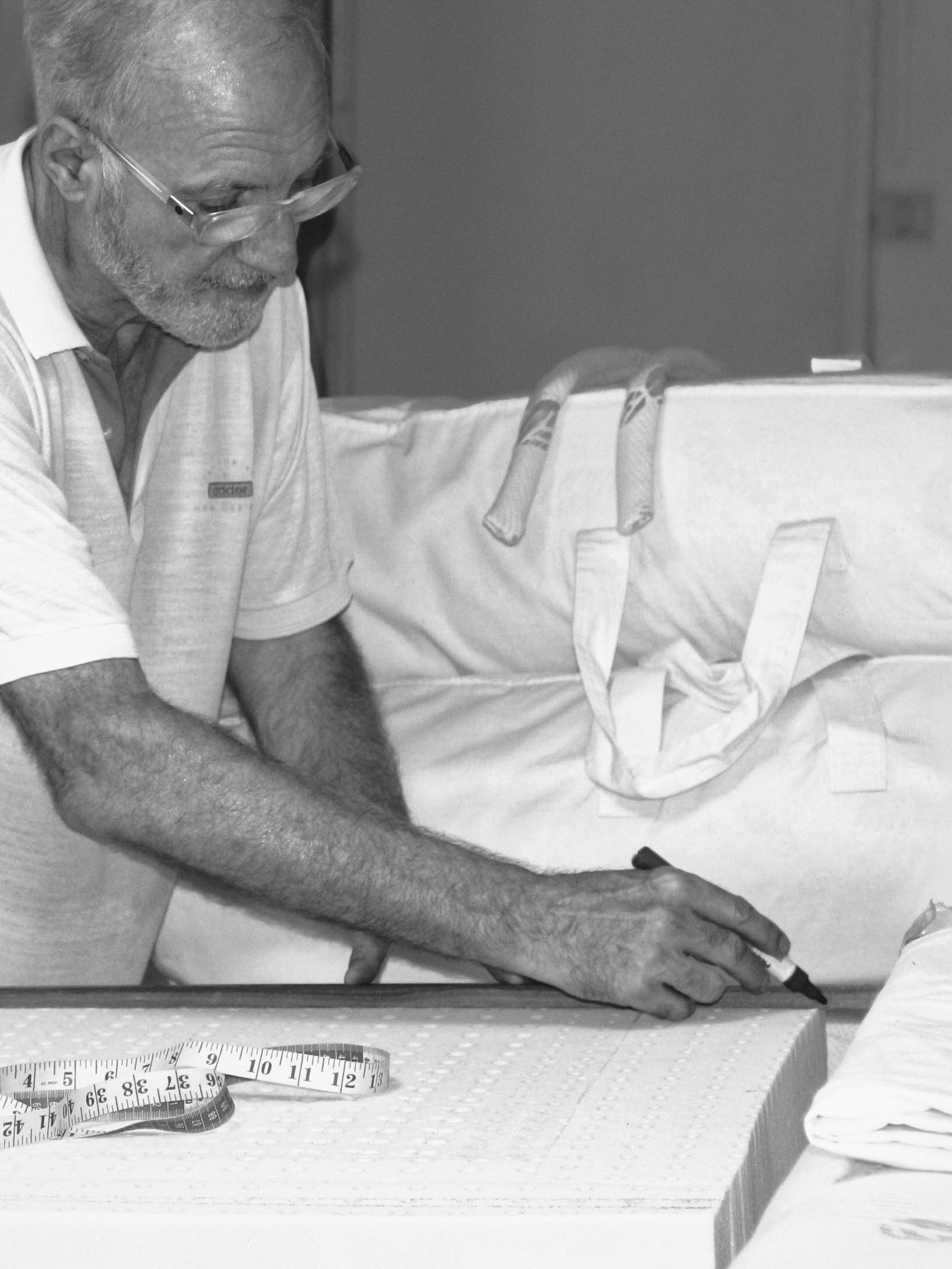 Dominique Bienfait in a white polo and glasses, leaning over a block of latex and drawing a line with a marker along a wooden rule, a tape measure in front of him.