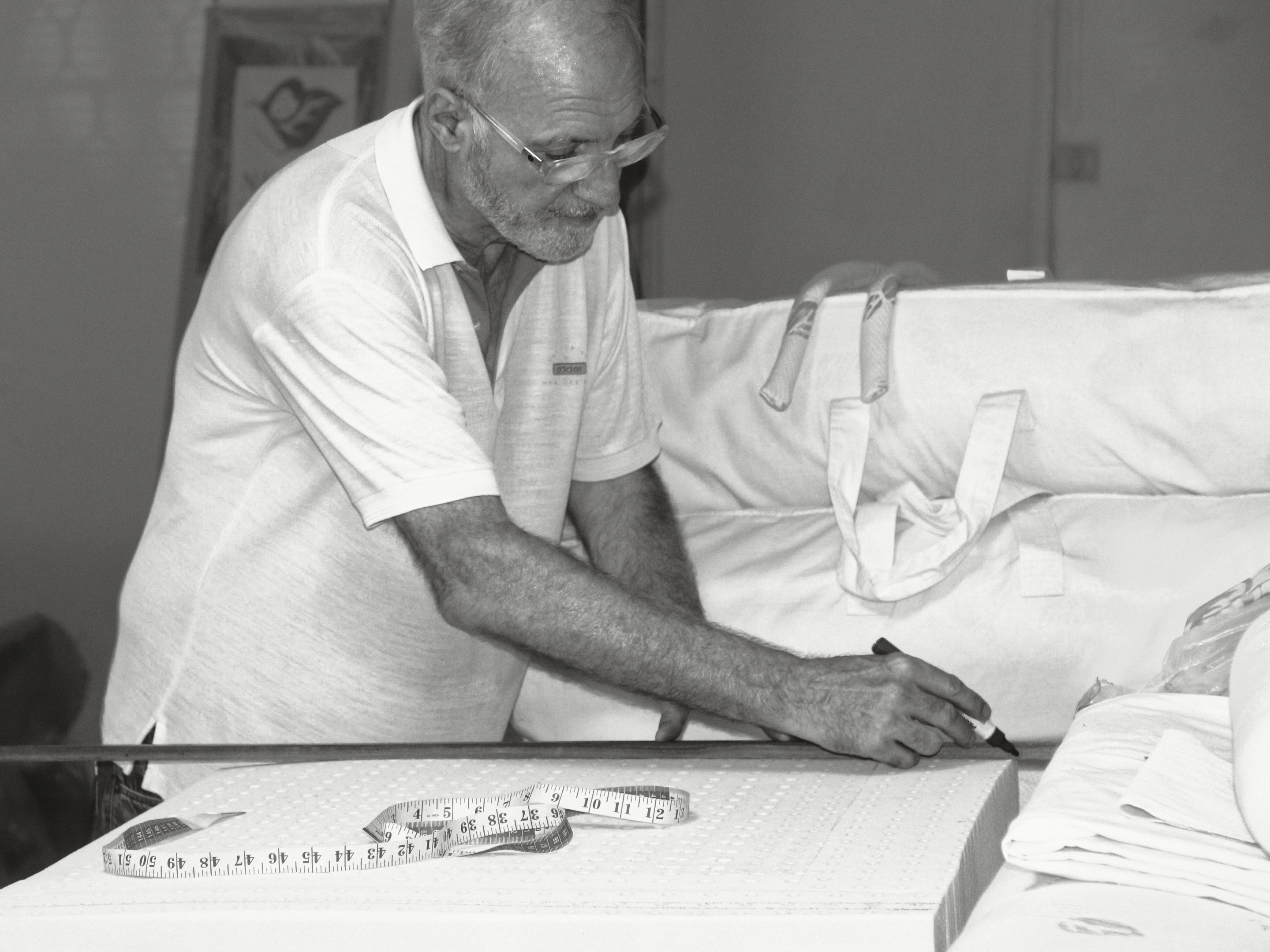 Dominique Bienfait in a white polo and glasses, bent over a block of latex on his work table, drawing a line with a marker along a wooden rule, a tape measure lying on the block and white cotton covers stacked behind him.