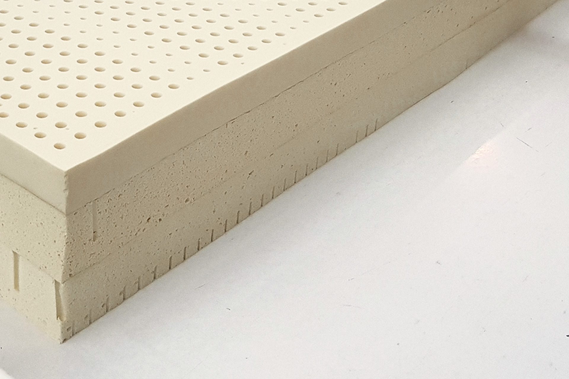 The near corner of a stack of three slabs of latex on a white table: the smooth perforated top, the open cells of the cut sides, a row of slits along the lower edge.