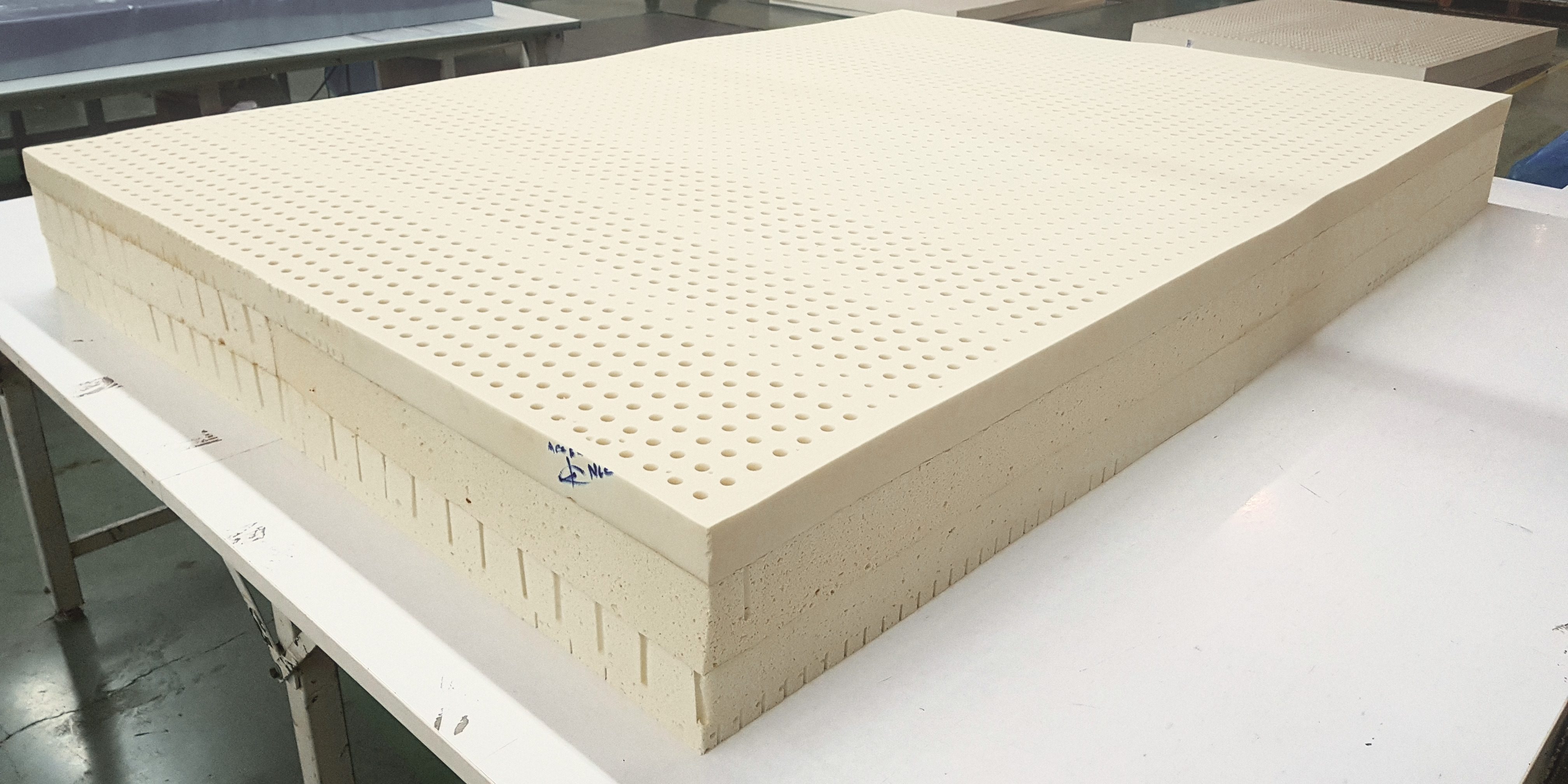 Three thick slabs of perforated latex stacked on a white table in a factory hall, the top slab covered in rows of pin holes, slits cut along the sides of the lower slabs.