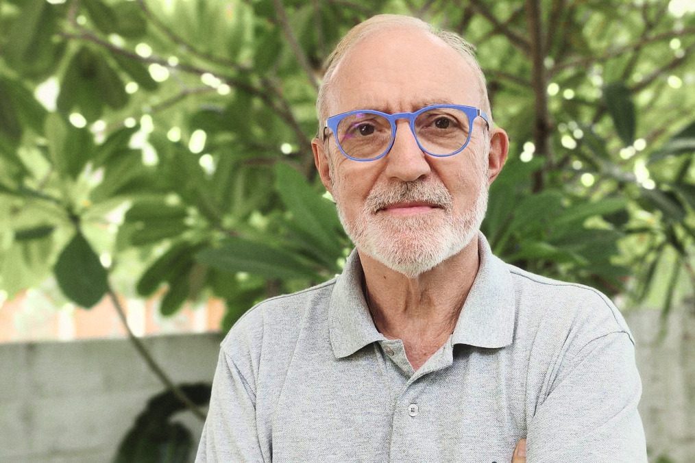 Dominique Bienfait, de près, en lunettes bleues et polo gris, des feuilles vertes derrière lui