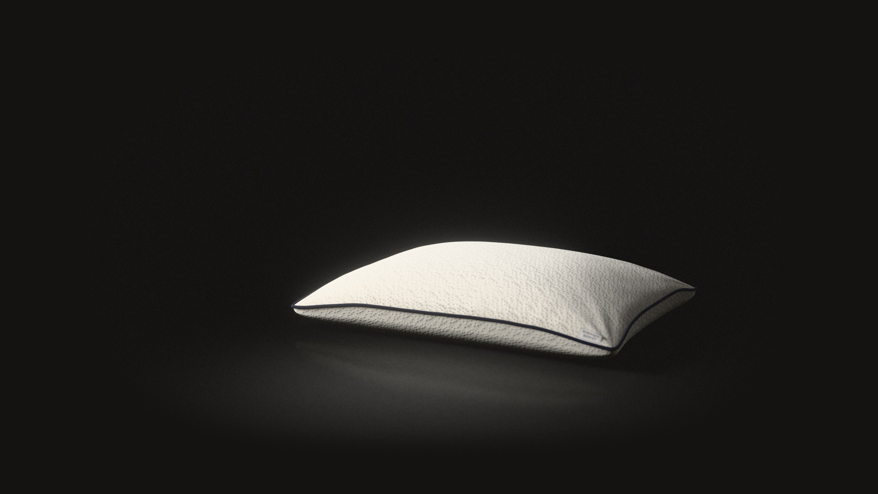 A pillow in a cream knit cover with dark piping and a small sewn label at its corner.