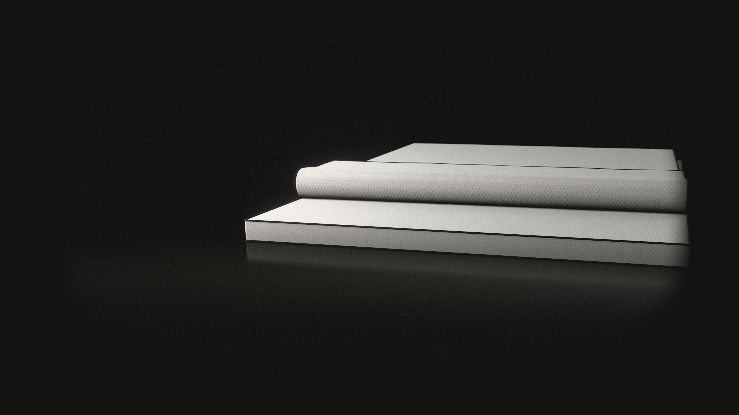 Two white latex toppers on a black floor, one lying on the other: the thicker one flat, the thinner one with its foot end folded back, each edged with black cord.