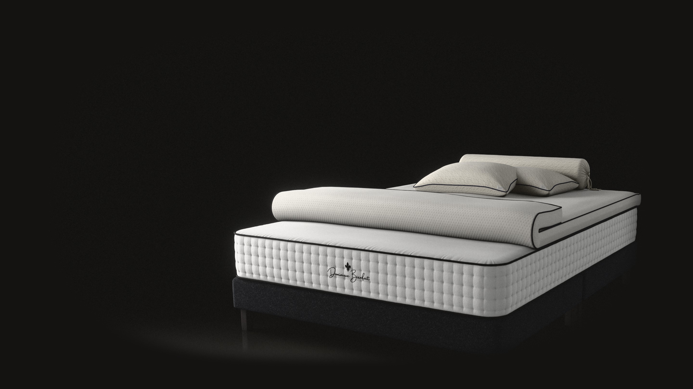 A bed in a black studio, seen from its foot: a white tufted latex mattress with the fleur-de-lis and Dominique Bienfait’s name on its foot end, standing on a low charcoal base. A white topper lies on it, its foot end folded back; two cream pillows and a bolster lie at the head.