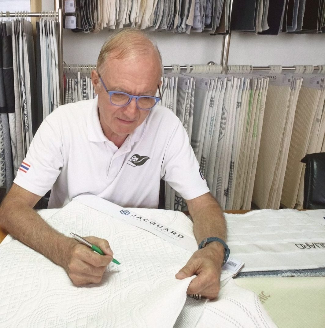Dominique Bienfait in a white polo and blue glasses at a desk, a green pen in his hand, marking a white quilted fabric, racks of hanging fabric samples behind him.