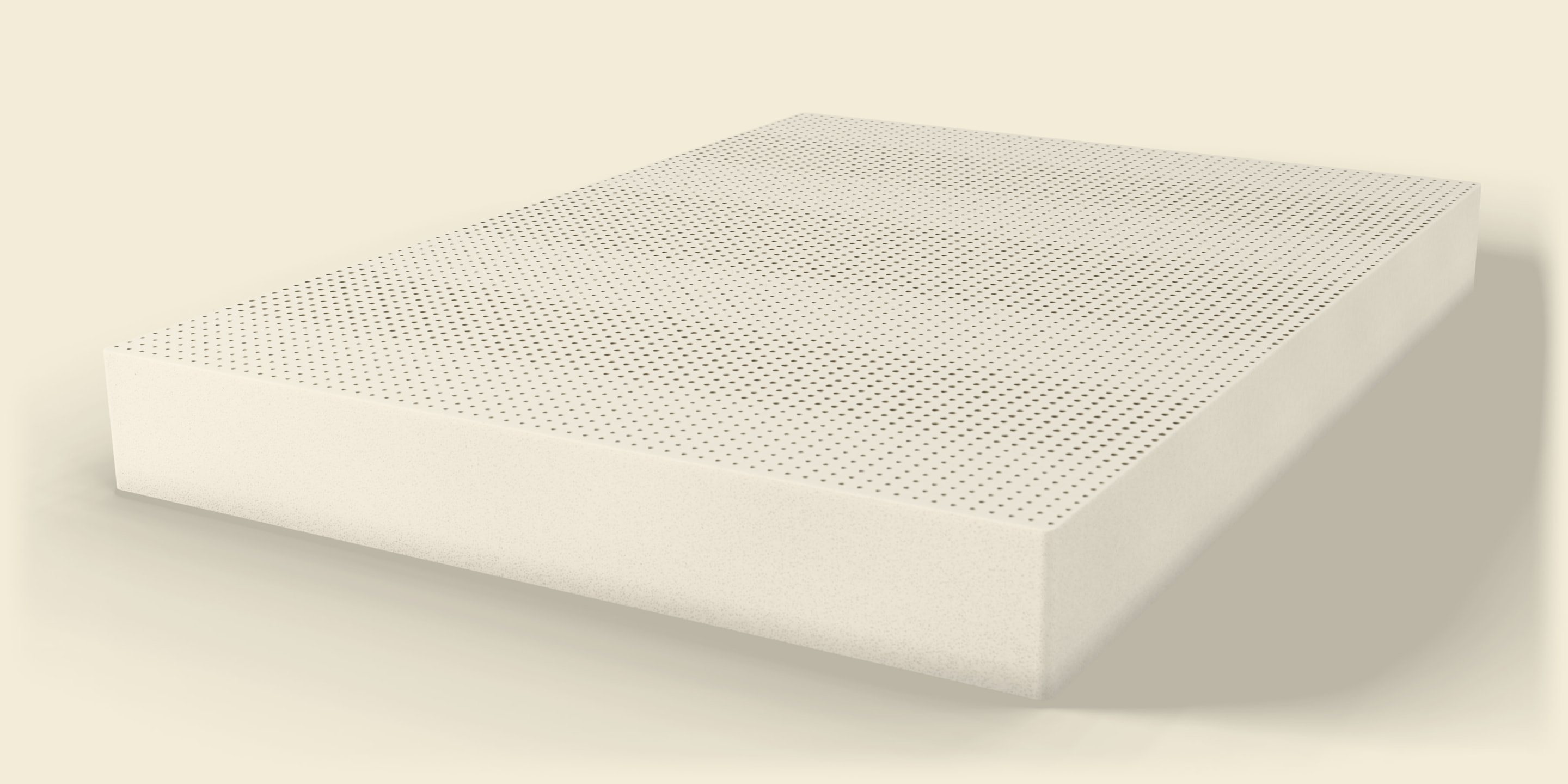 A block of cream natural latex in daylight, seen low from one corner, its top covered in rows of pin holes, its soft shadow on the floor.