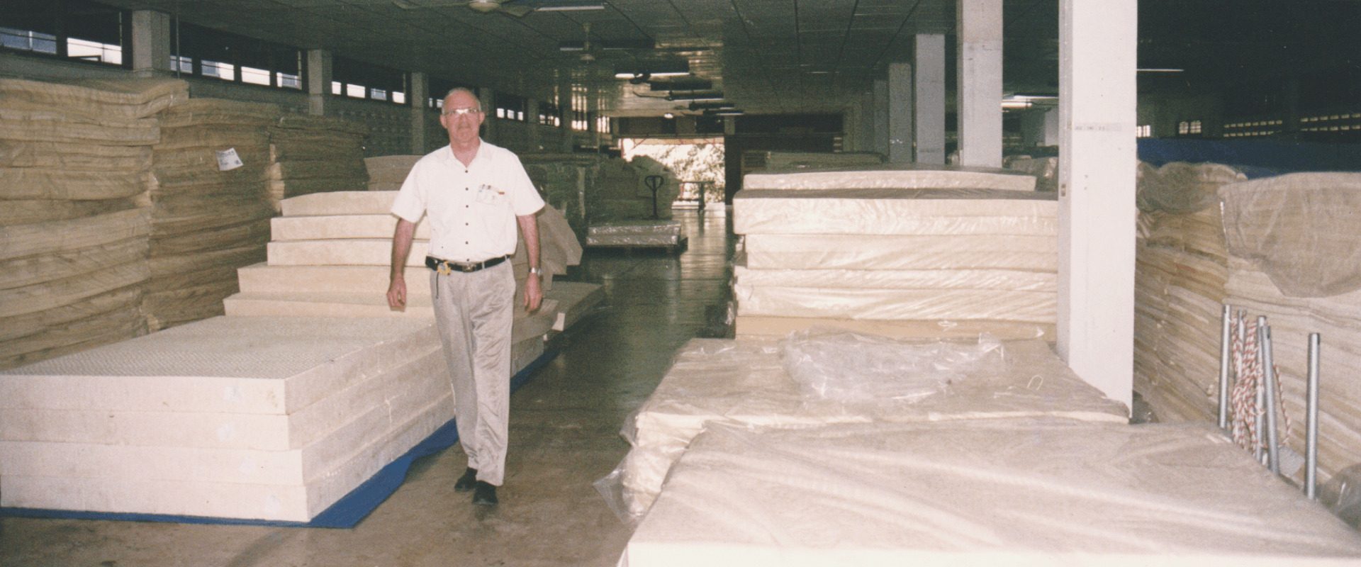 An old colour print: Dominique Bienfait in a white shirt walking down an aisle of a large factory hall between tall stacks of cream latex slabs.