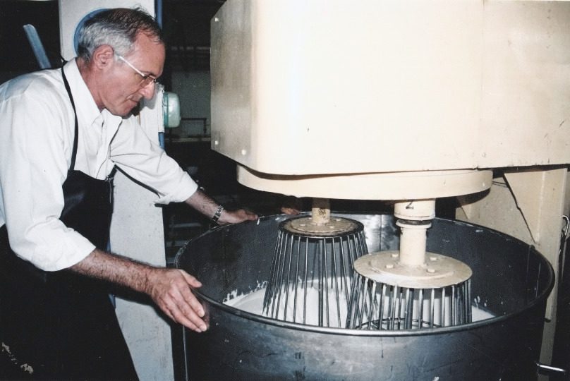 An old colour print: a man in a white shirt and a black apron leans over a steel bowl in which two cage whisks beat white latex, under the cream housing of a foaming machine.