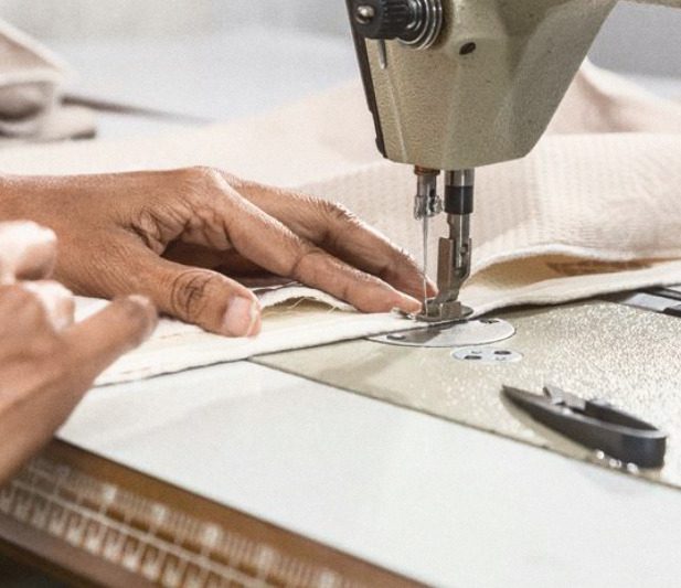 Two hands feeding the folded edge of a white knitted panel under the needle of a sewing machine, a pair of thread snips on the table.