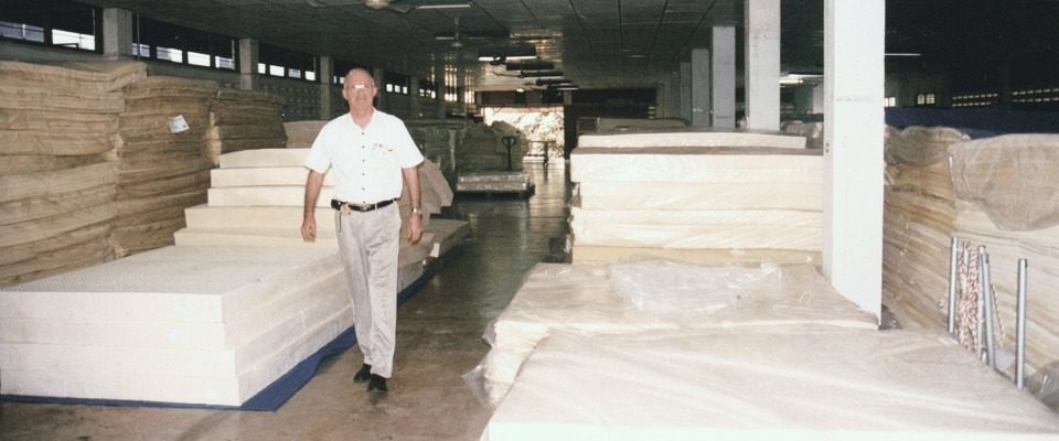 An old colour print: Dominique Bienfait in a white shirt walks down an aisle between tall stacks of latex in a large factory hall.