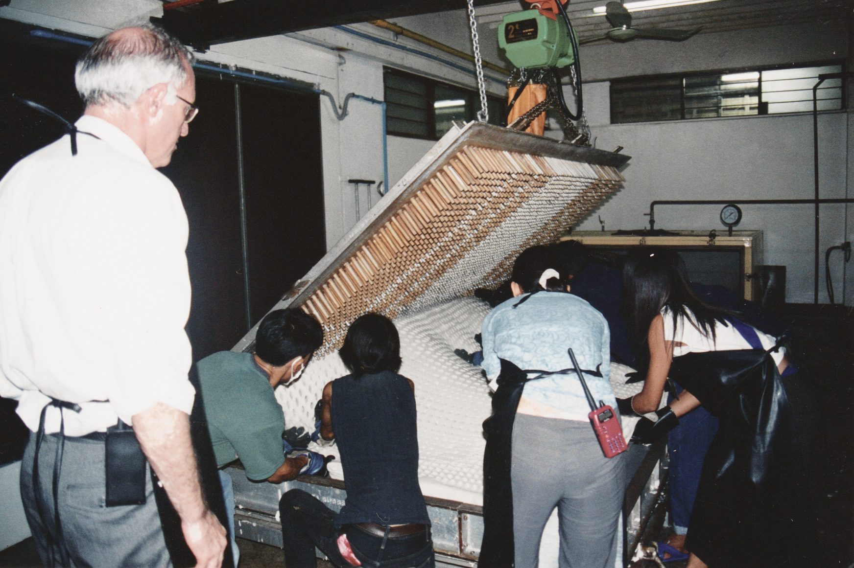 An old colour print: the lid of a mould, covered in rows of long pins, lifted on a hoist while four workers ease a white latex core out from under it, Dominique Bienfait watching from the left.