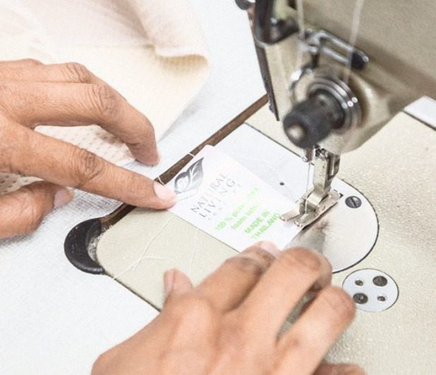 Two hands holding a woven label flat under the foot of a sewing machine, on white knitted cloth.